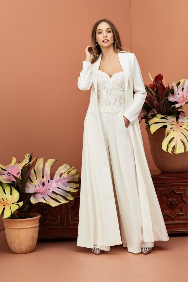 a woman in a white dress and jacket standing next to a vase with flowers on it