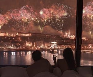 two people sitting on a bed watching fireworks go off in the night sky over a city