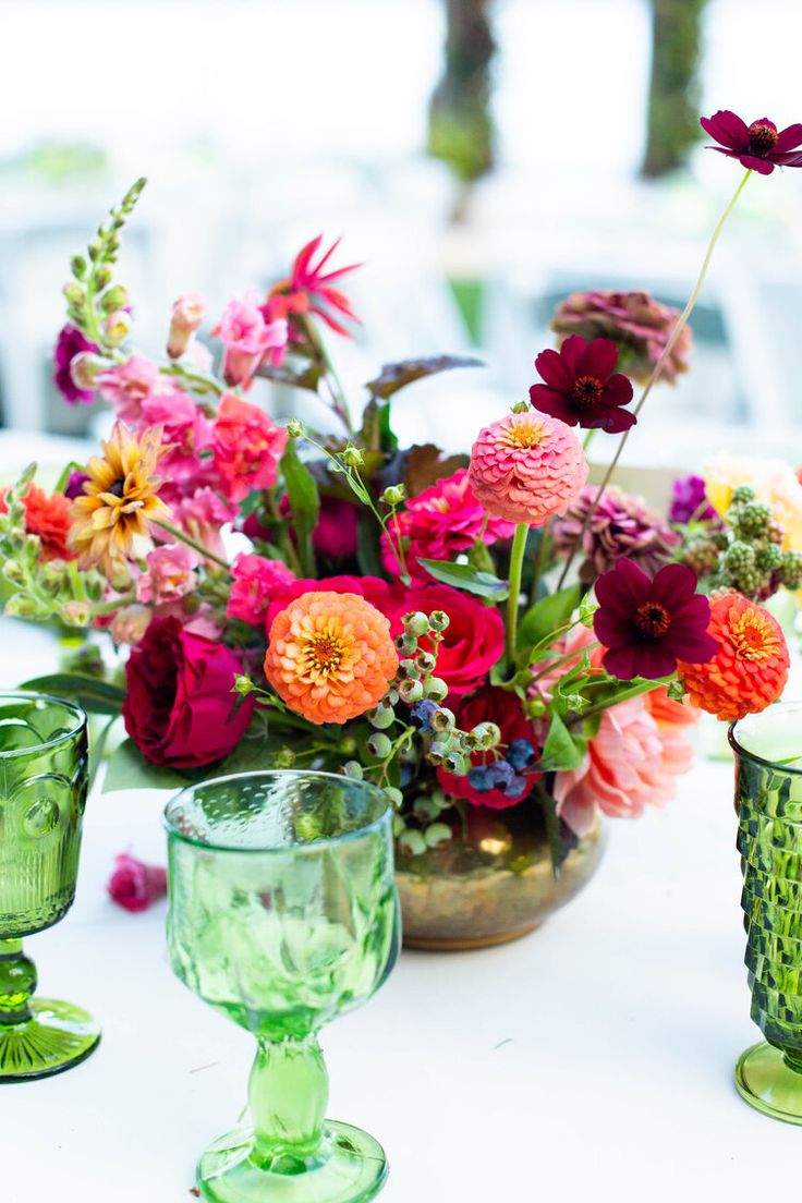 colorful flowers are in vases and glasses on a white table with green glassware