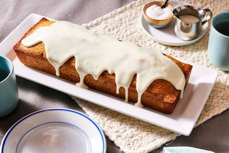 a loaf of bread with white icing sitting on a plate next to cups and saucers