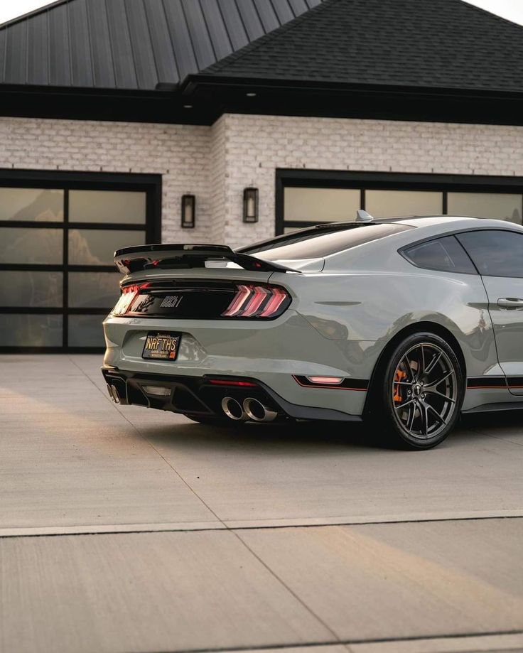 the rear end of a gray mustang parked in front of a house with two garage doors