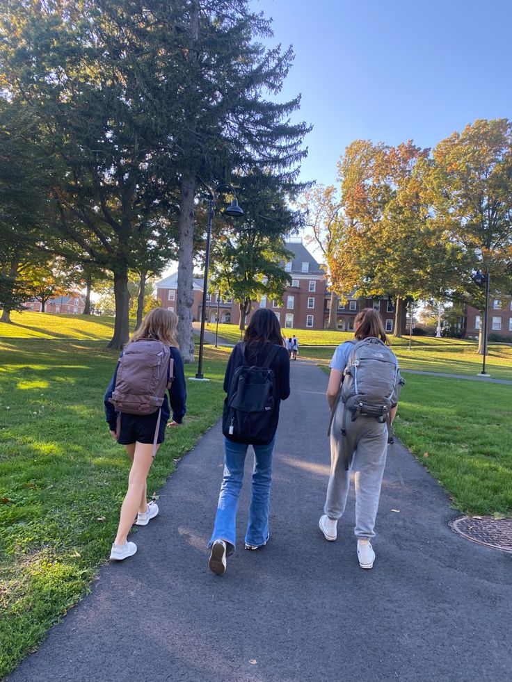 three girls walking down a path in the park with backpacks on their backs and one girl looking at her cell phone