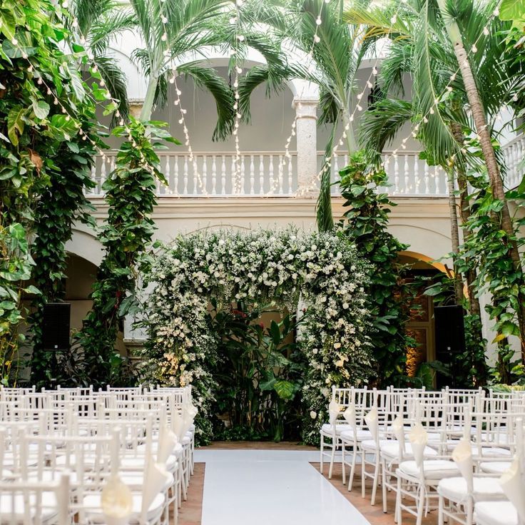 an indoor wedding venue with white chairs and greenery on the walls, surrounded by palm trees