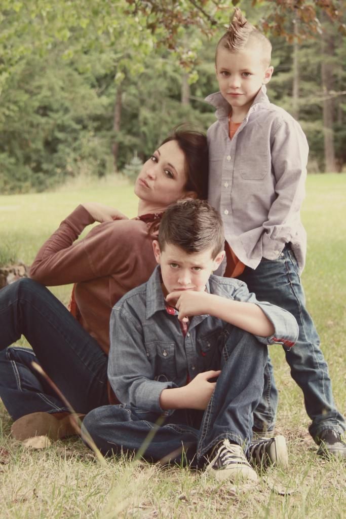 a woman and two boys sitting in the grass under a tree with their arms around each other