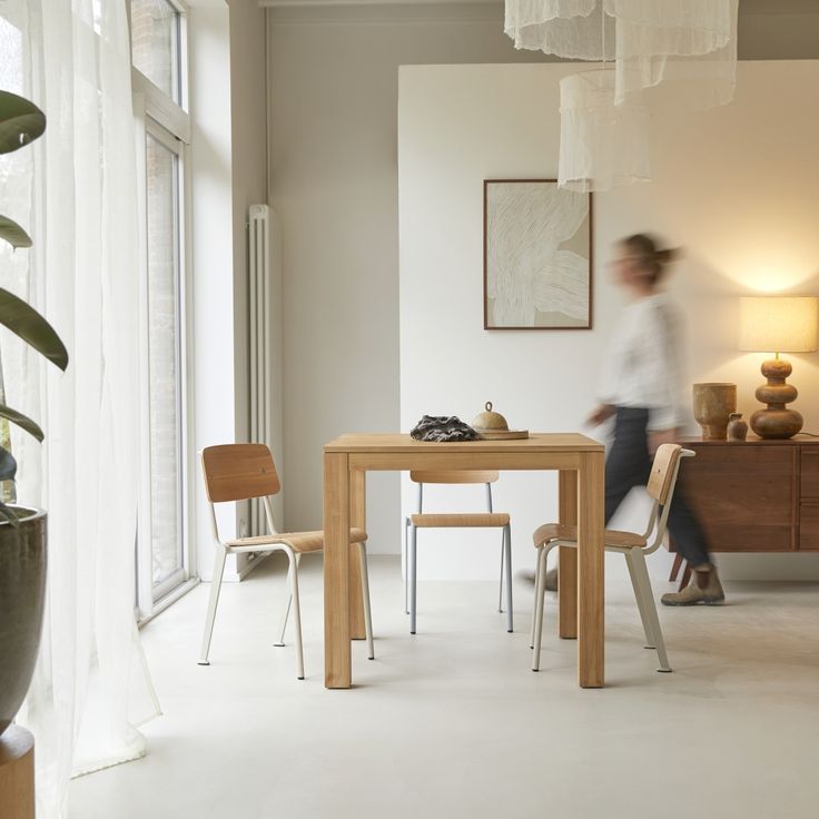 a woman walking past a wooden table in a room with white walls and flooring