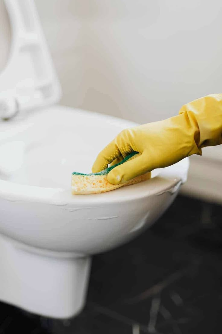 a person in yellow gloves cleaning a white toilet bowl with a sponge and green rag