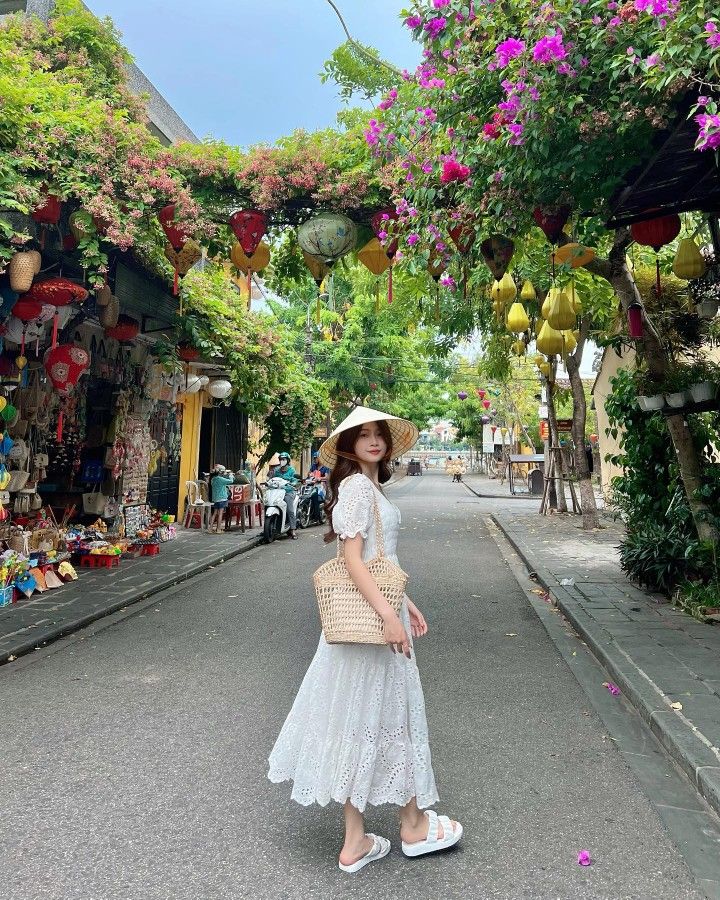 a woman in a white dress and hat is standing on the street with flowers hanging above her head