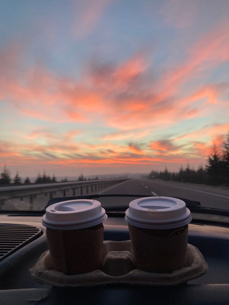 two cups of coffee sit on the dashboard of a car as the sun goes down