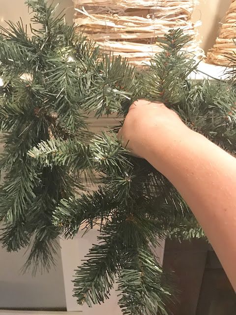 a person holding a fake pine branch in front of a basket on the wall with wicker baskets behind it