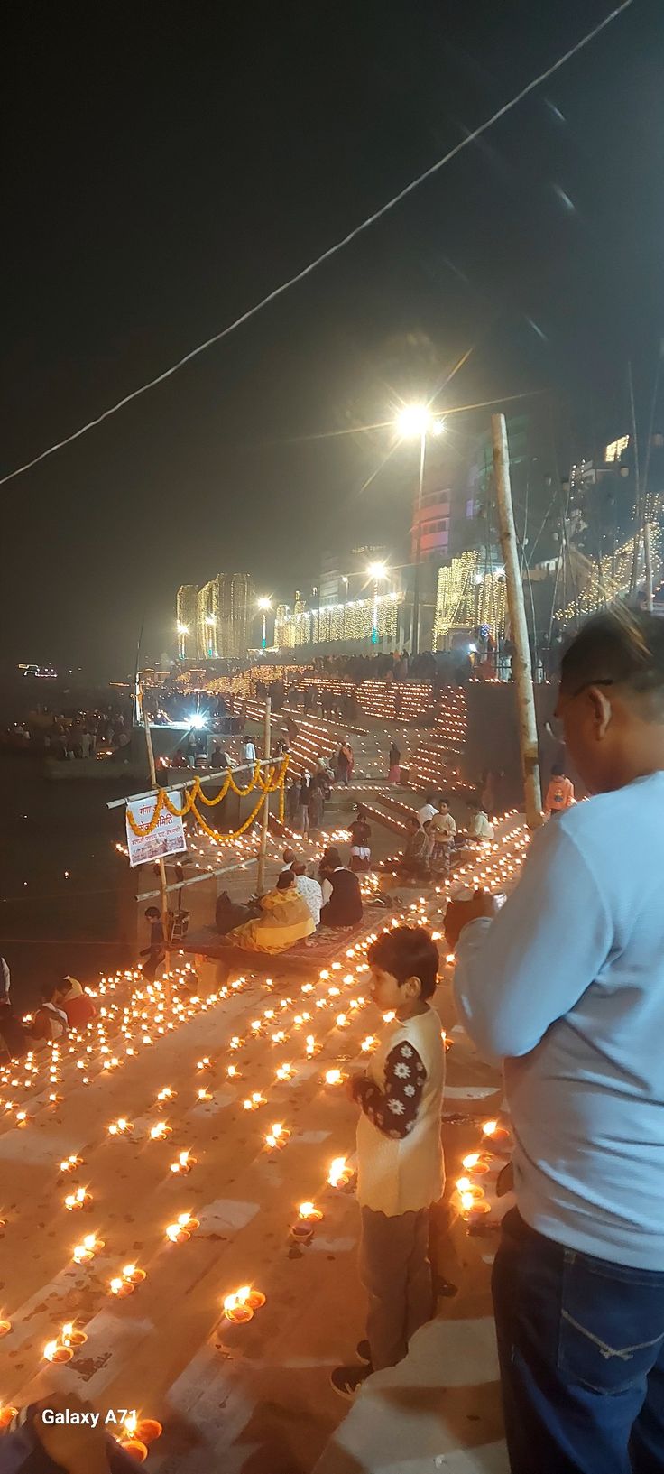 many people are standing around with candles on the ground and lights in the water behind them