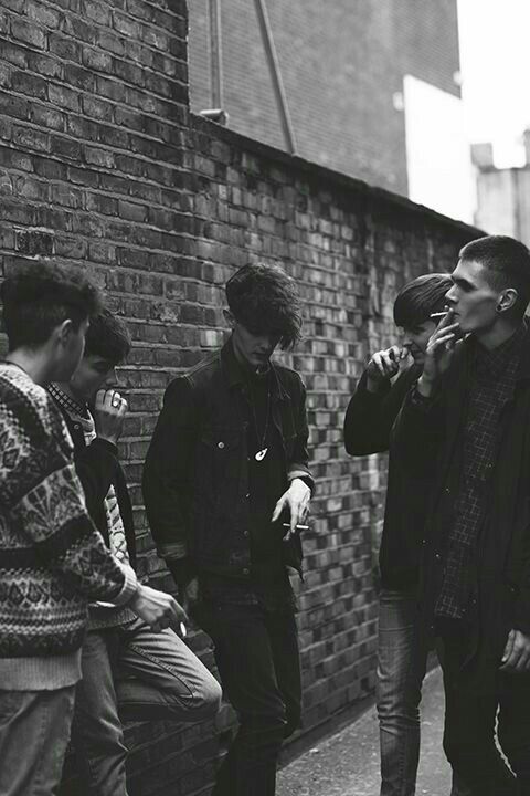 four young men standing next to a brick wall talking on their cell phones while one looks at his phone