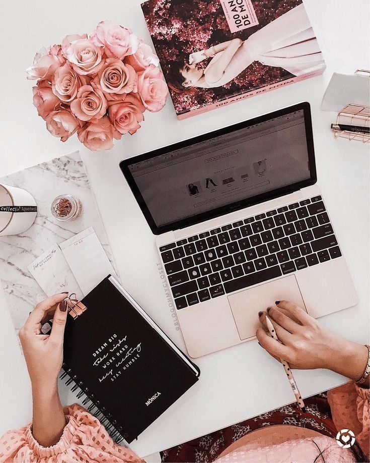 a woman sitting at a desk with a laptop and notebook in front of her, surrounded by pink flowers