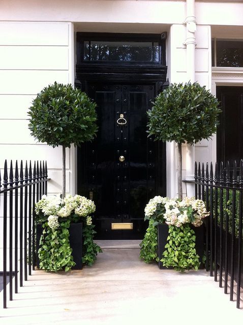 two black planters with white flowers in front of a door on a house's sidewalk