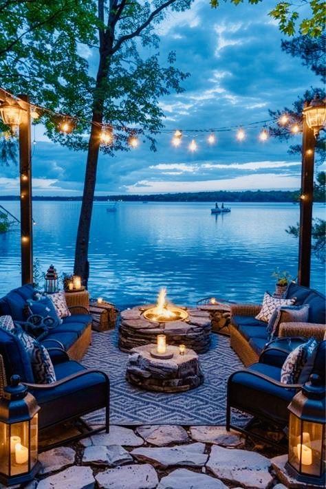 an outdoor seating area with fire pit, chairs and lights on the water's edge