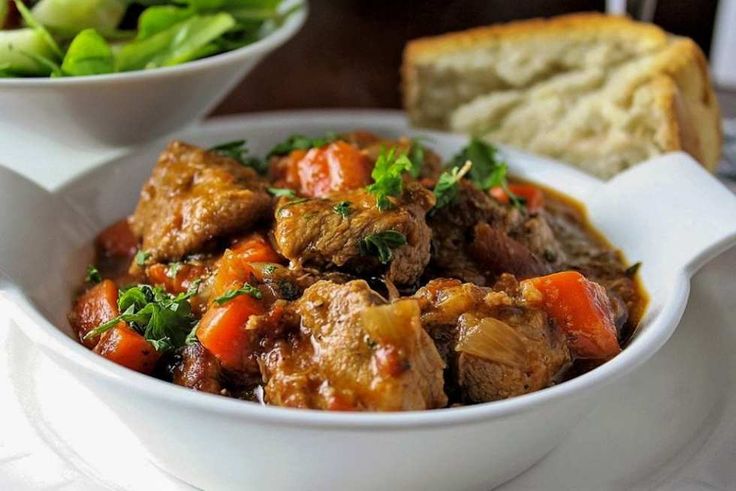 a white bowl filled with meat and vegetables next to a slice of bread on a table