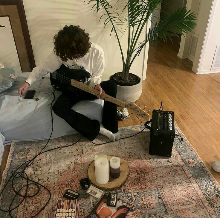 a person sitting on the floor with a guitar and some other items around them in front of a potted plant