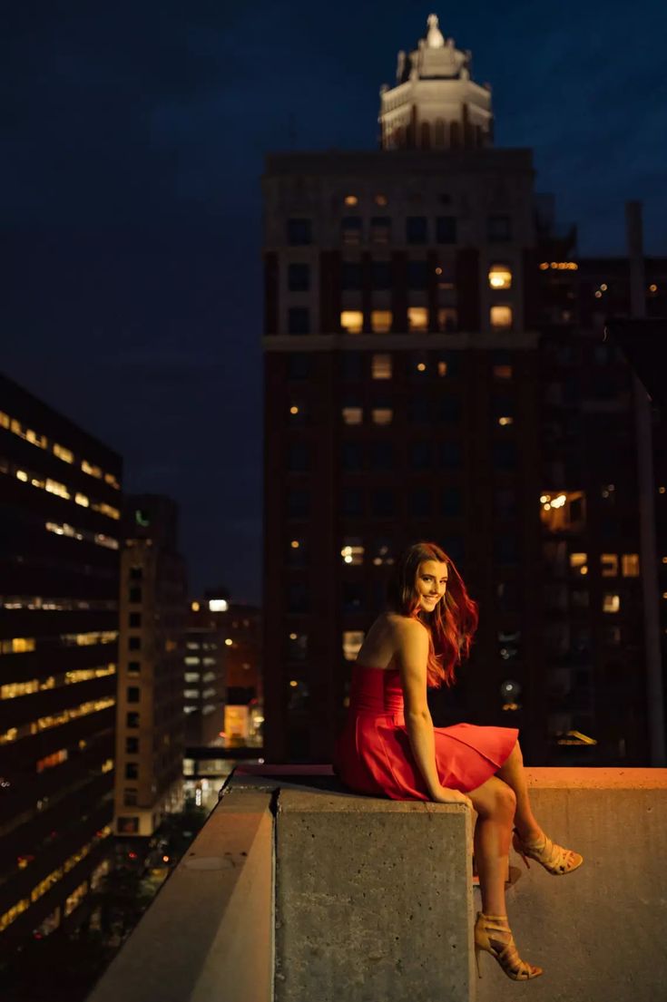 a woman in a red dress is sitting on a ledge at night, with buildings in the background