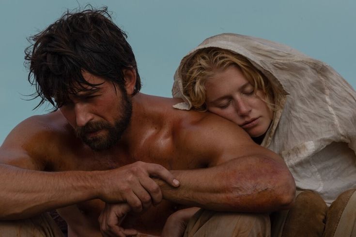 a shirtless man and woman sitting next to each other on the ground in front of a blue sky