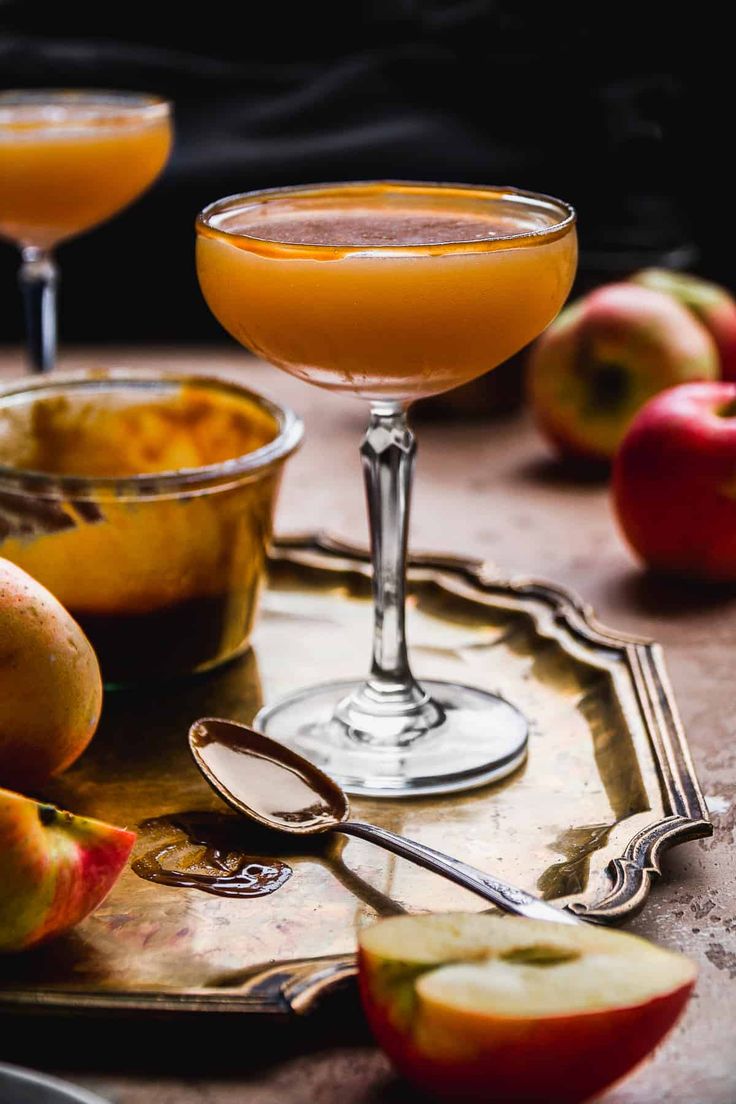 two glasses filled with orange juice next to apples and spoons on a tray in front of them