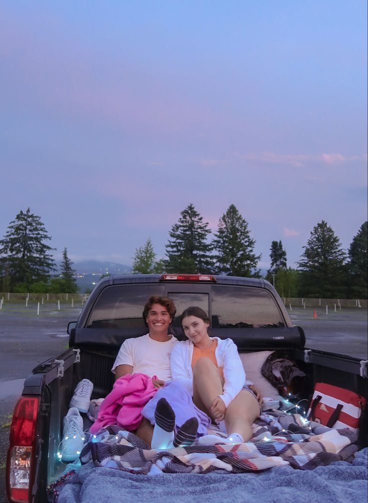 a man and woman sitting in the back of a pick up truck on a blanket