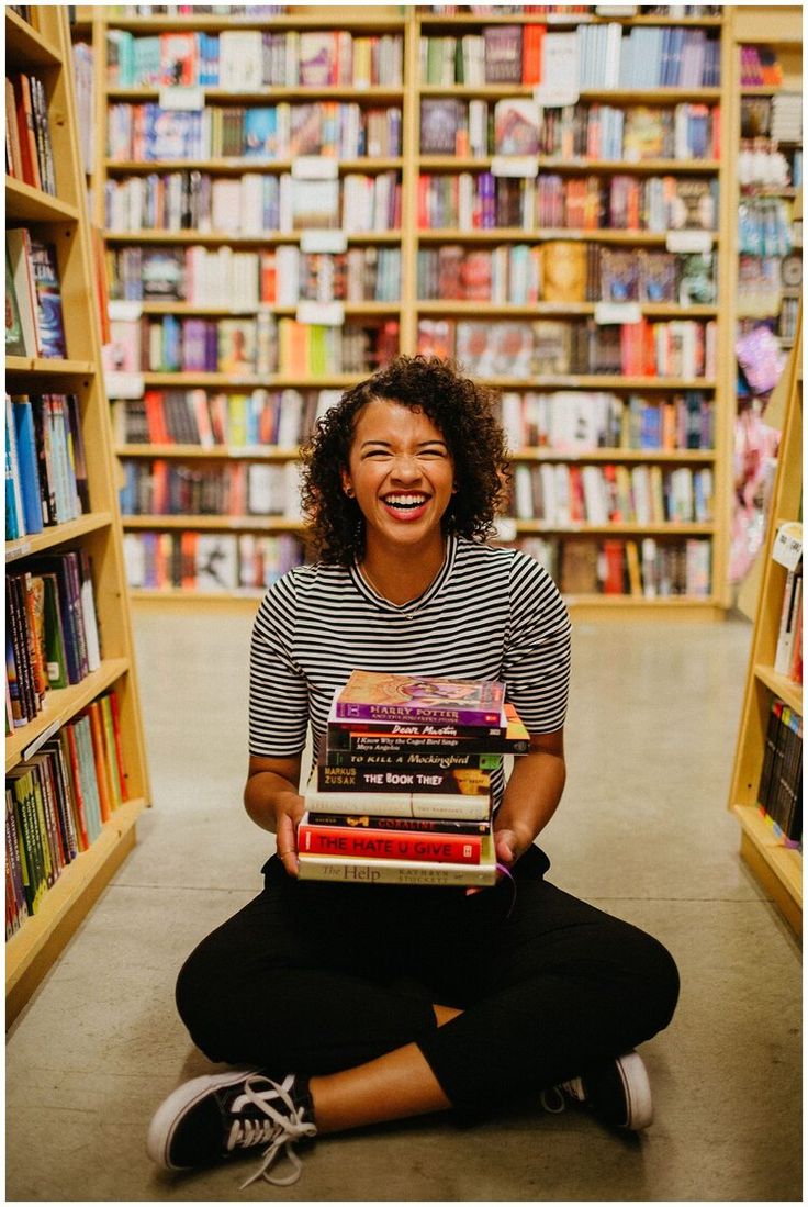 a woman sitting on the floor in front of a bookshelf holding a stack of books