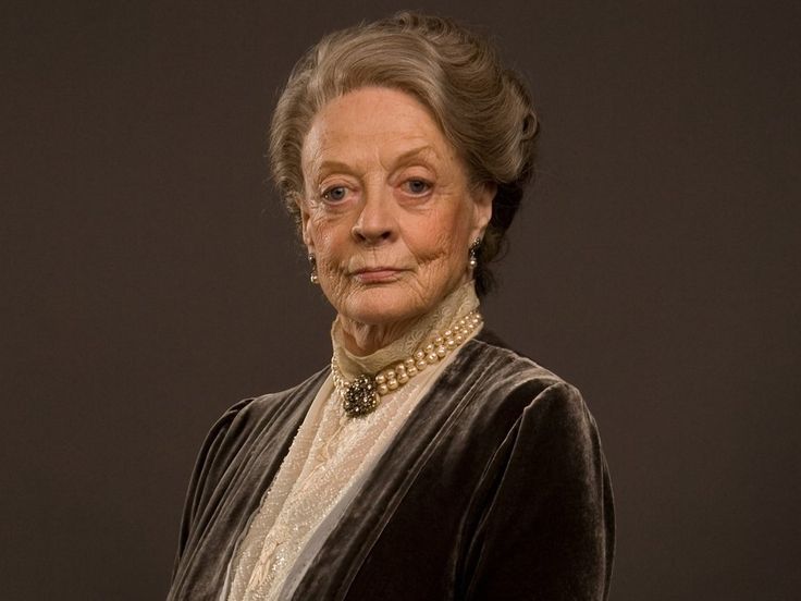 an older woman in a black dress and pearls on her head is looking at the camera