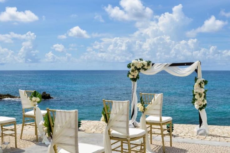 an outdoor wedding setup with white chairs and flowers on the aisle, overlooking the ocean