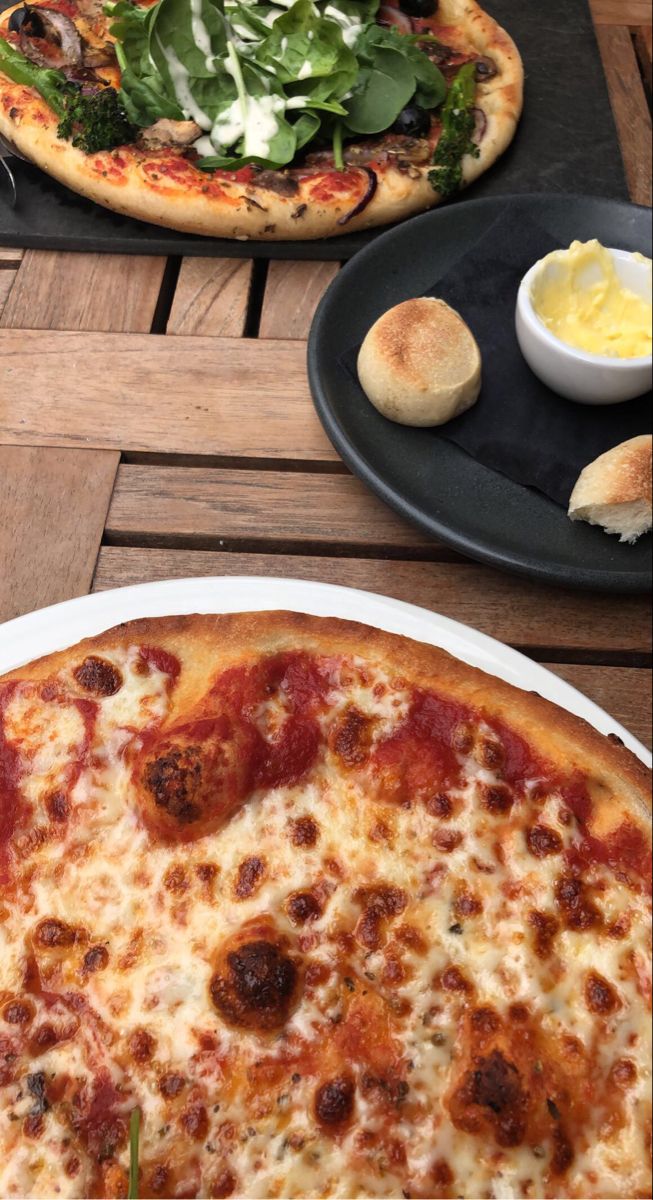 two pizzas sitting on top of a wooden table next to plates with food in them