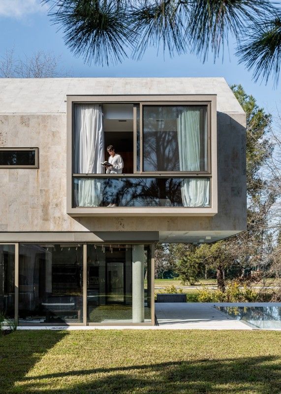 a person standing in the window of a modern house overlooking a pool and trees on a sunny day