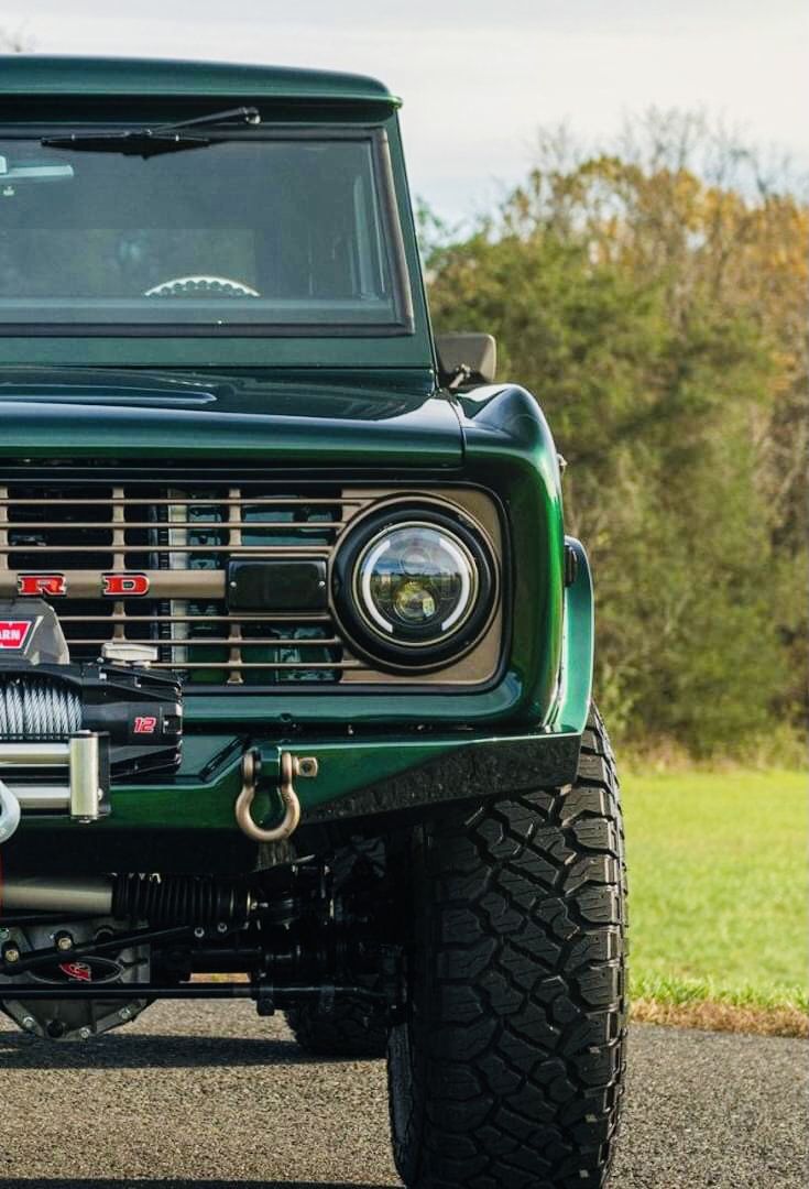 the front end of a green jeep parked in a parking lot next to grass and trees