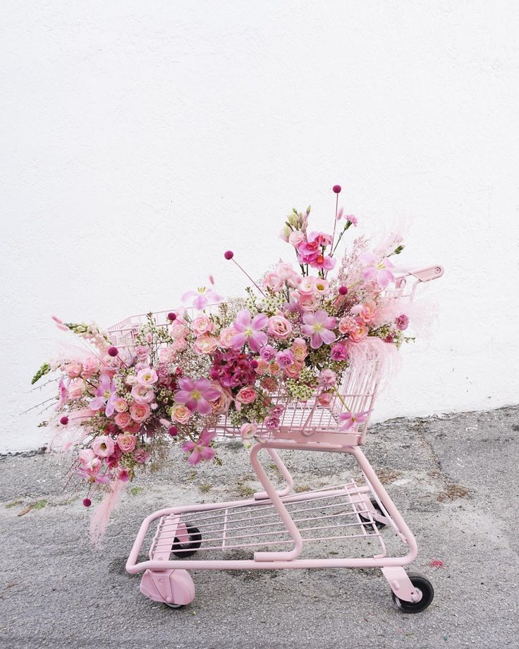 a pink shopping cart with flowers in it on the ground next to a white wall