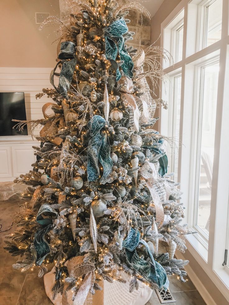 a christmas tree with blue and silver ornaments in a living room next to a large window