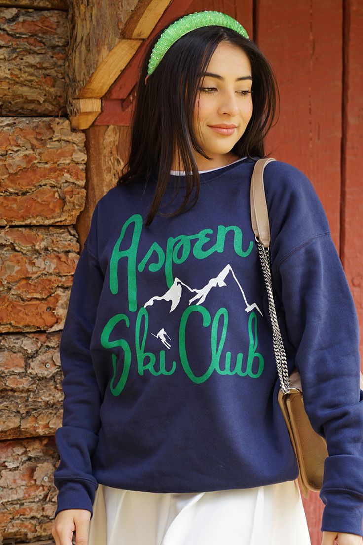 a woman wearing a blue sweatshirt with green lettering on the front and back, standing in front of a brick wall