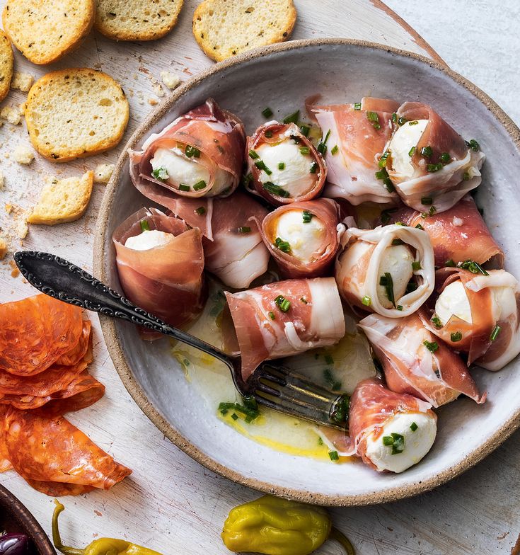 a bowl filled with food next to crackers and pepperoni on top of a table