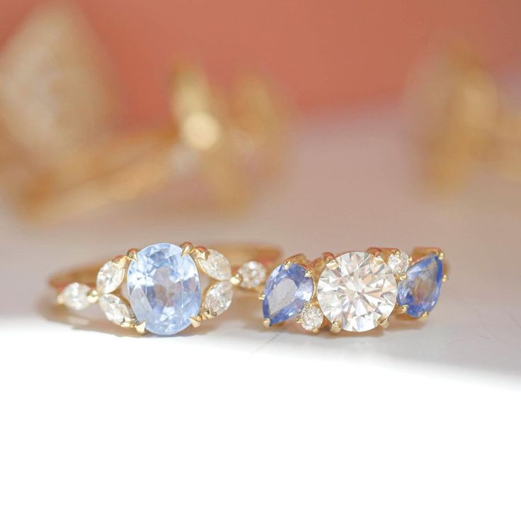 two gold rings with blue and white stones on the sides, one has an oval shaped diamond