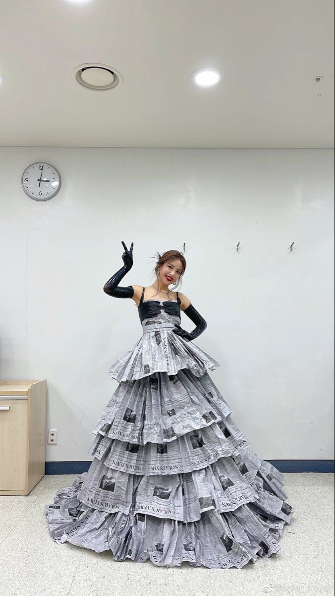 a woman in a dress made out of newspapers standing next to a wall with a clock on it