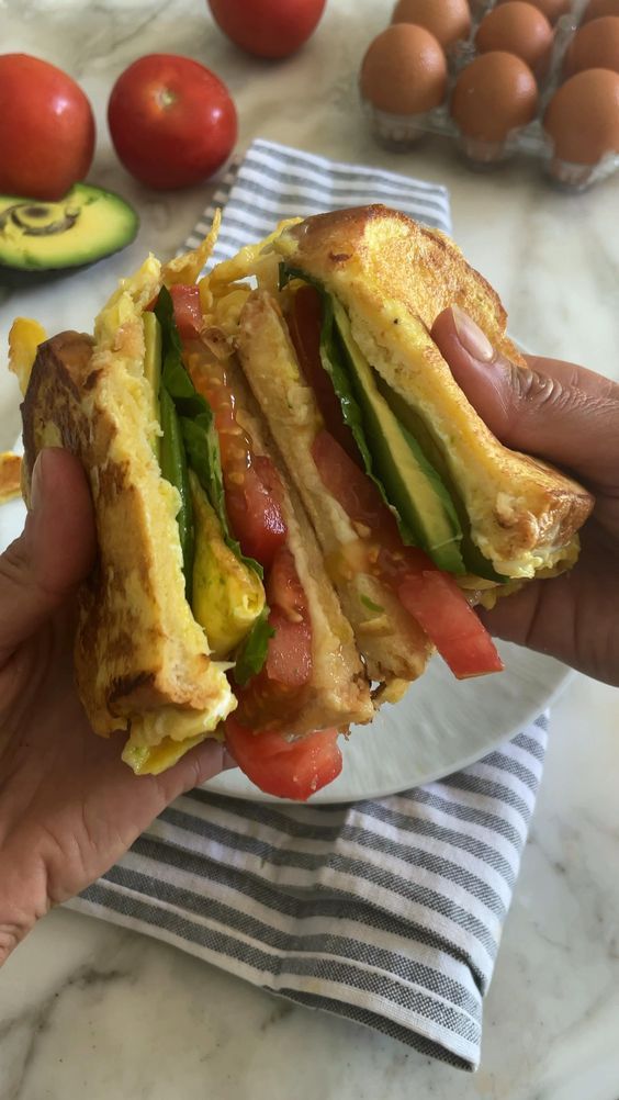 a person holding up a sandwich with tomatoes, avocado and egg in the background