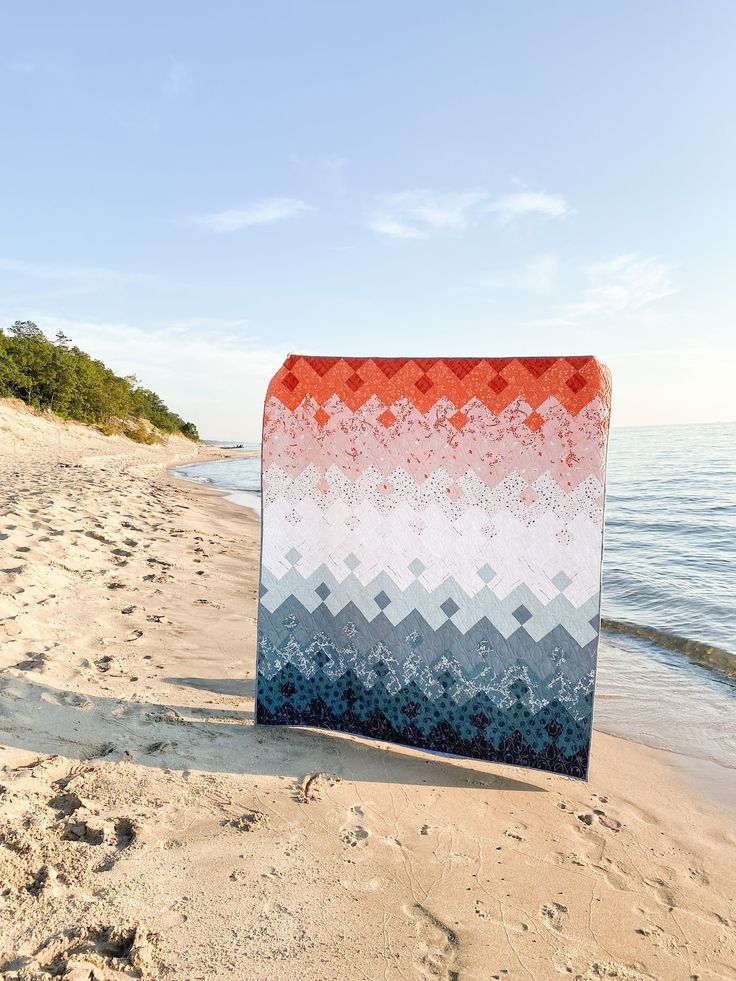 an orange, white and blue blanket sitting on top of a sandy beach