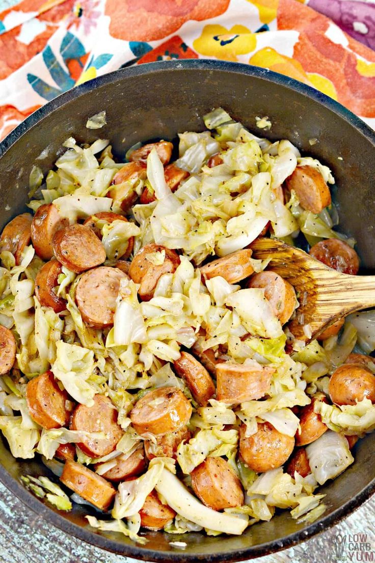 a pan filled with cabbage and sausages on top of a colorful table cloth next to a wooden spoon