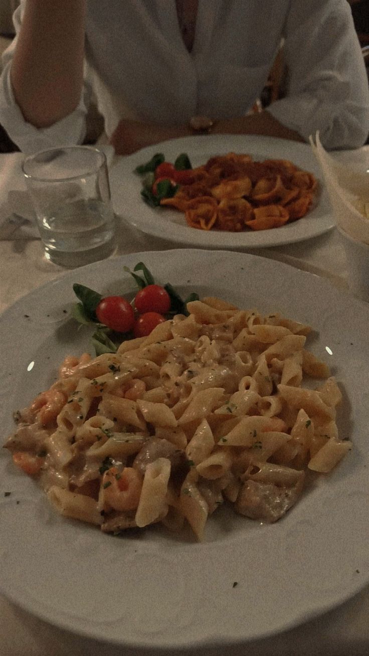 a woman sitting at a table with two plates of food on it, one plate has pasta and the other is vegetables