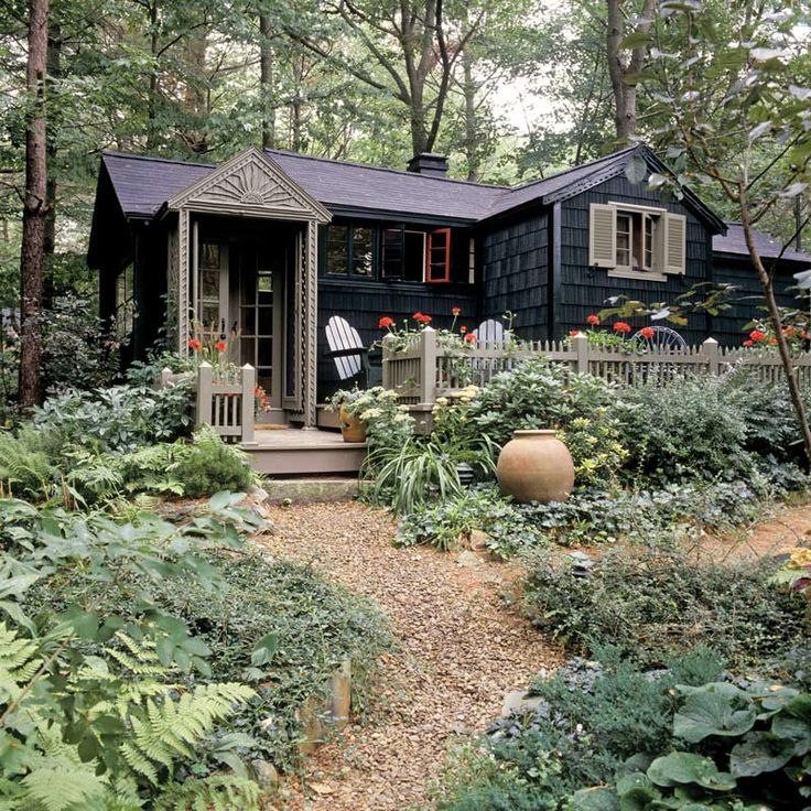 a small black house surrounded by trees and plants in the woods with chairs on the front porch