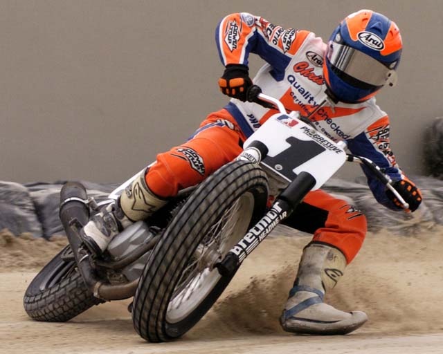 a man riding on the back of a motorcycle down a dirt road in an orange and white suit