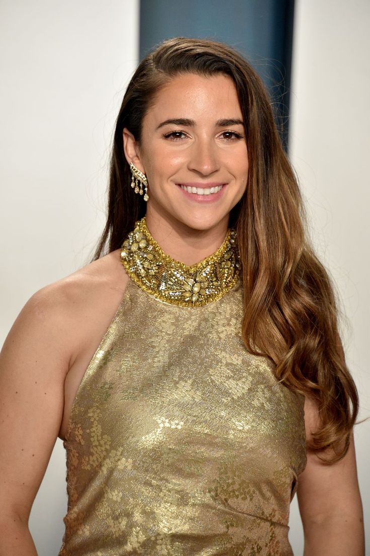 a woman with long hair wearing a gold dress and statement necklace smiling at the camera