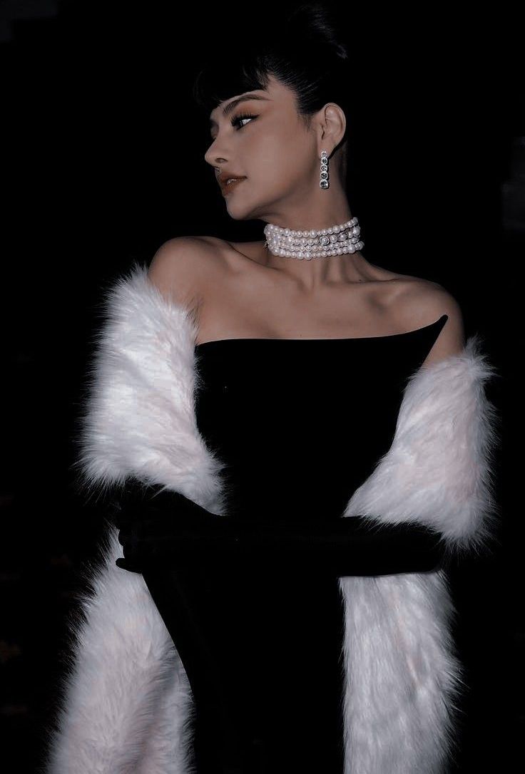 a woman in a black dress and white fur stole with jewels on her neck, standing against a dark background