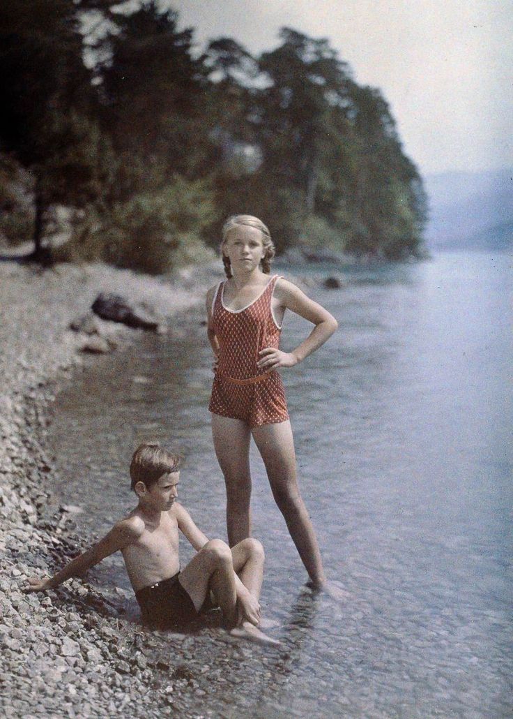 two children are sitting in the water near some rocks and trees, one is wearing a bathing suit