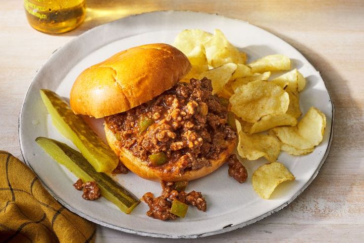 a sloppy joe sandwich with pickles and chips on a plate