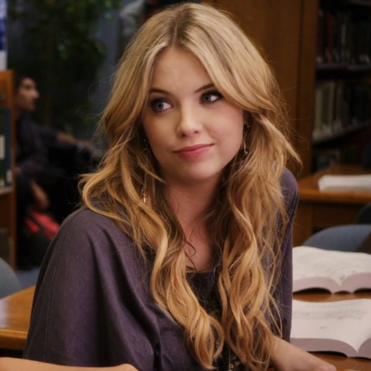 a woman with long blonde hair sitting in front of a book shelf and looking at the camera