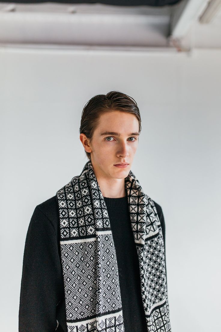 a man wearing a black and white scarf standing in front of a white wall, looking at the camera