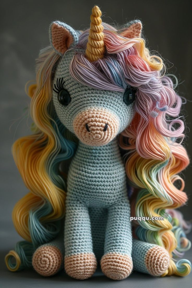 a crocheted stuffed unicorn sitting on top of a gray surface with rainbow hair