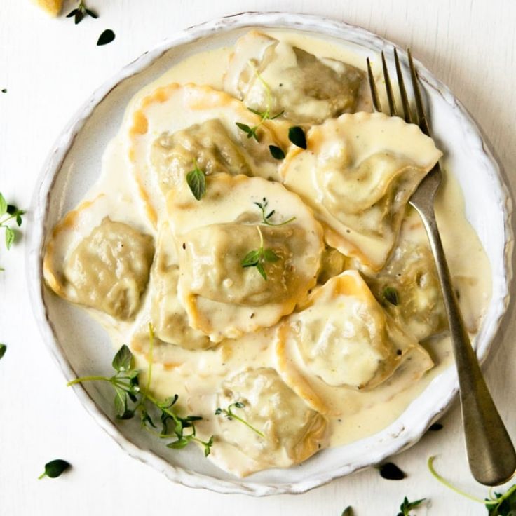 a white plate topped with ravioli next to a fork and green leafy garnish